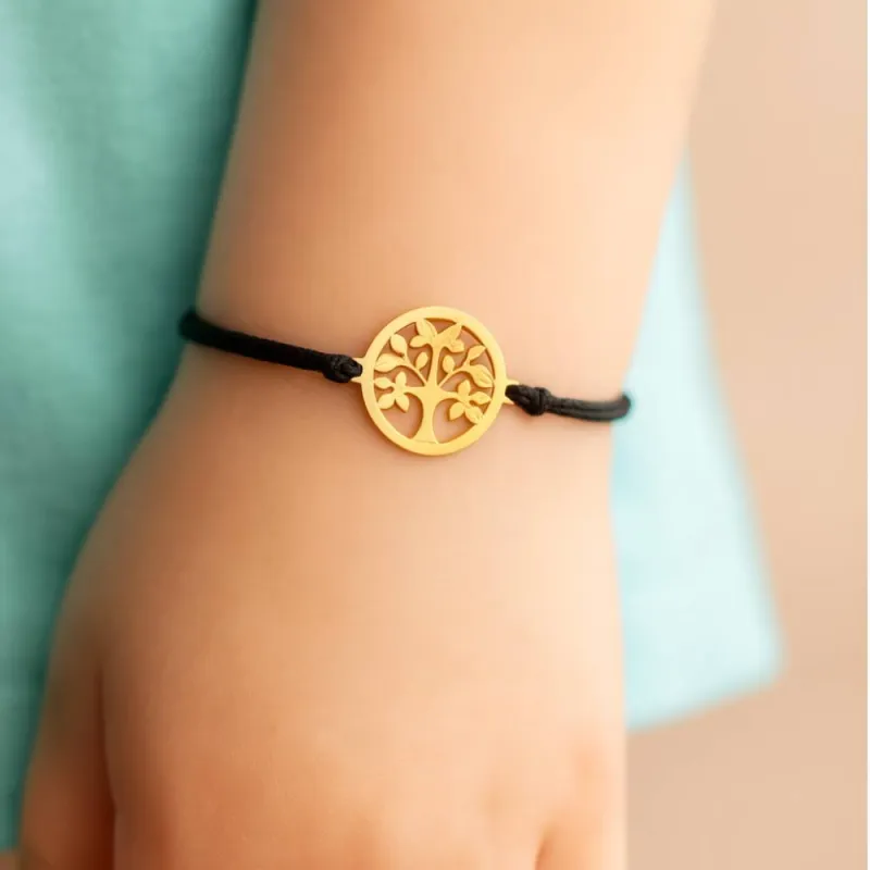 Bracelet en or avec pendentif arbre de vie porté sur cordon noir, un bijou moderne et symbolique souvent offert comme cadeau de communion.