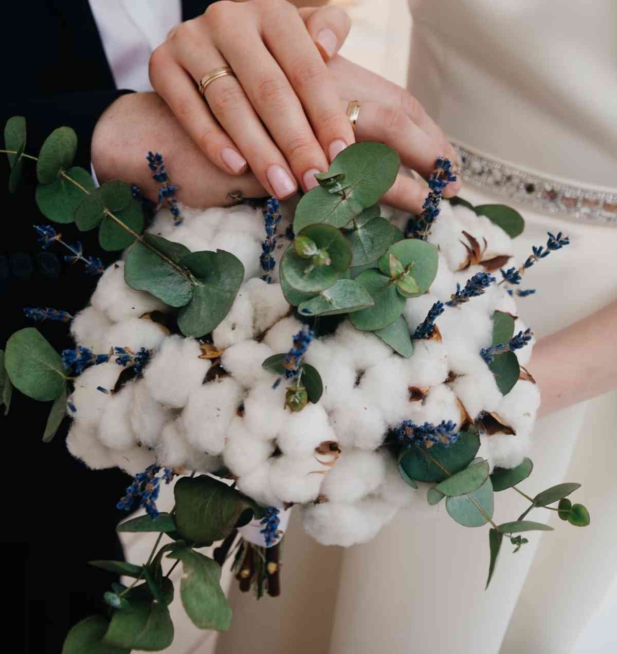 Gros plan sur deux mains enlacées portant des alliances en or, posées sur un bouquet de coton et d’eucalyptus ; l’image illustre la signification des noces de coton et l’engagement d’un couple après une première année d’union.