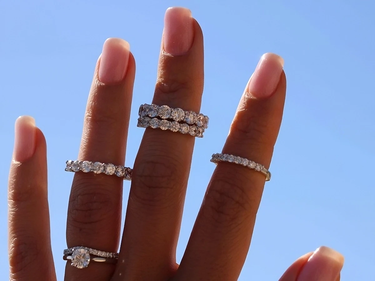 Collection de bagues en diamant sous le soleil Main ornée de multiples bagues en diamant étincelant, capturée sous un ciel bleu clair, mettant en valeur l'éclat des bijoux.