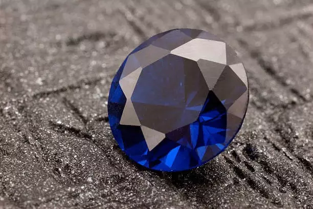 Gros plan d’un diamant bleu taille ronde posé sur une surface noire texturée, mettant en valeur sa brillance unique.