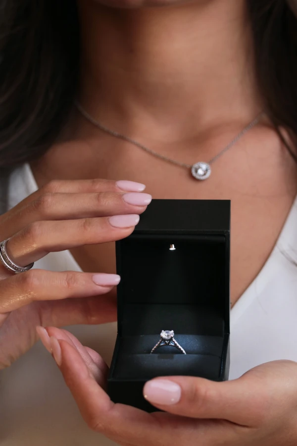 Boîte à bijoux ouverte contenant une bague de fiançailles solitaire en diamant avec une lumière intégrée, tenue par une femme portant un collier élégant.