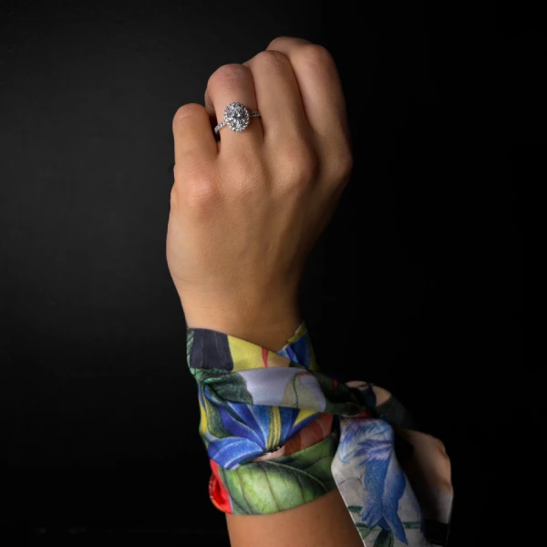 Mano llevando un anillo de diamante engastado, acompañado de una bufanda Mini Twill colorida atada alrededor de la muñeca, sobre fondo negro