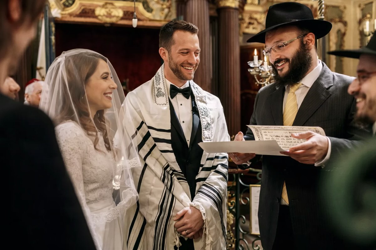 Pareja celebrando una boda judía en la sinagoga mientras el rabino lee la ketubá, momento solemne previo al ritual de romper el vaso.