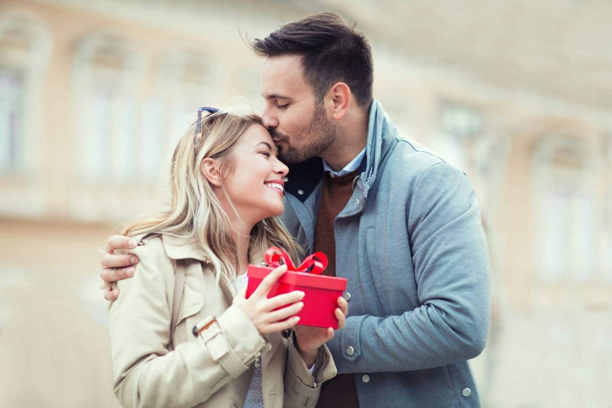 Une femme sourit en tenant un cadeau rouge tandis que son mari l’embrasse sur le front, scène romantique marquant la célébration des 13 ans de mariage, ou noces de muguet.