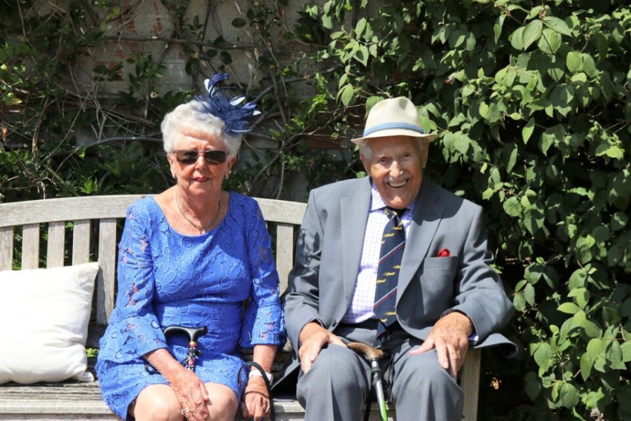 Un couple âgé souriant, la femme en robe bleu roi avec chapeau assorti et l’homme en costume gris avec chapeau clair, assis sur un banc en bois dans un jardin, célébrant la solidité et la longévité de leurs noces de chêne après 80 ans de mariage.
