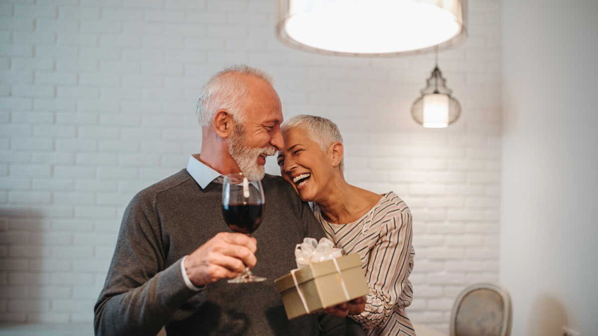 Un couple senior souriant et complice, partageant un verre de vin et un cadeau, lors de la célébration de leurs 34 ans de mariage, symbole des noces d’ambre et de leur amour durable.