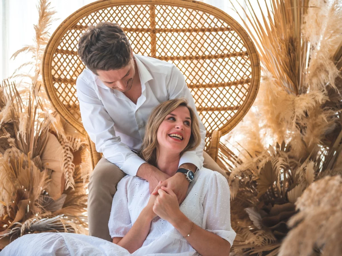 Pareja celebrando su aniversario de boda en un entorno natural y bohemio - Celinni Una pareja sonriente, vestida de blanco, celebra su aniversario de boda en un ambiente acogedor con elementos naturales, simbolizando el amor y la complicidad después de dos años de matrimonio (bodas de cuero).