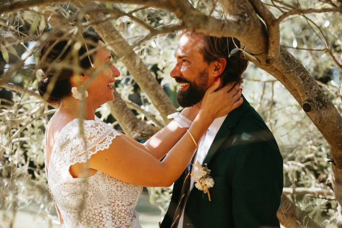Un couple de mariés souriant se tient tendrement sous les branches d’un arbre, dans un moment romantique capturé le jour de leur mariage.