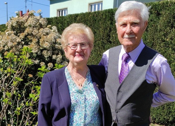 Un couple âgé élégant, la femme en veste bleue et l’homme en gilet gris avec cravate violette, pose ensemble dans un jardin ensoleillé, symbole de la force et de la longévité des noces de chêne après 80 ans de mariage.