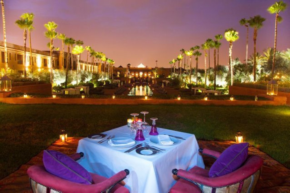 Table dressée pour deux dans un jardin illuminé de Marrakech, entourée de palmiers et lanternes, cadre luxueux pour une demande en mariage inoubliable.