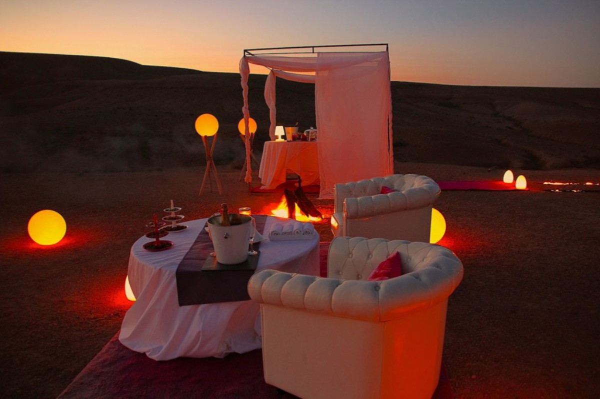 Table élégante dressée au coucher du soleil dans le désert d’Agafay, ambiance feutrée avec fauteuils blancs et bougies, parfaite pour une demande en mariage magique à Marrakech.