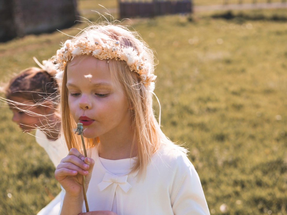 Petite fille en tenue blanche et couronne de fleurs soufflant sur un pissenlit, illustrant l’innocence et la douceur d’une journée de première communion.