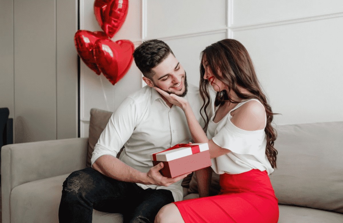 Femme offrant un coffret cadeau rouge à son compagnon, assis dans un salon décoré de ballons cœur ; scène inspirante pour un présent symbolique célébrant le premier anniversaire de mariage (noces de coton).