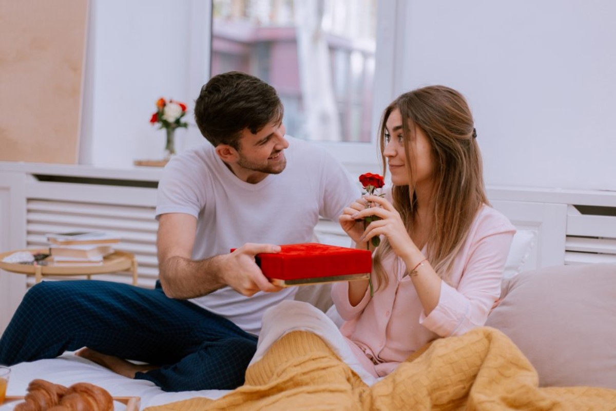 Un homme offre un cadeau et une rose rouge à une femme souriante au réveil, illustrant un moment tendre et romantique pour souhaiter un joyeux anniversaire à sa compagne.