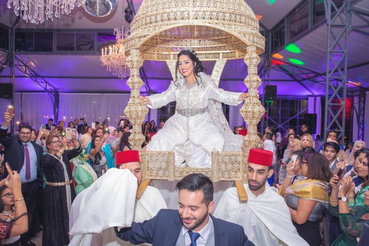 عروس مغربية محمولة في العمارية الذهبية – مراسم تقليدية عروس متألقة بقفطان أبيض ومجوهرات لامعة، جالسة في عمارية ذهبية يحملها رجال يرتدون أزياء تقليدية، وسط تصفيق الضيوف.