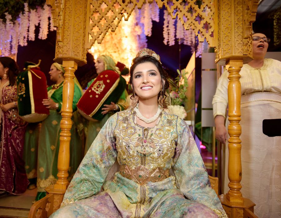 عروس مبتسمة في العمارية الذهبية – زفاف مغربي احتفالي عروس شابة مشرقة ترتدي تكشيطة ملوّنة، جالسة داخل عمارية منحوتة من الخشب، محاطة بنساء بملابس تقليدية وأجواء موسيقية مغربية احتفالية.