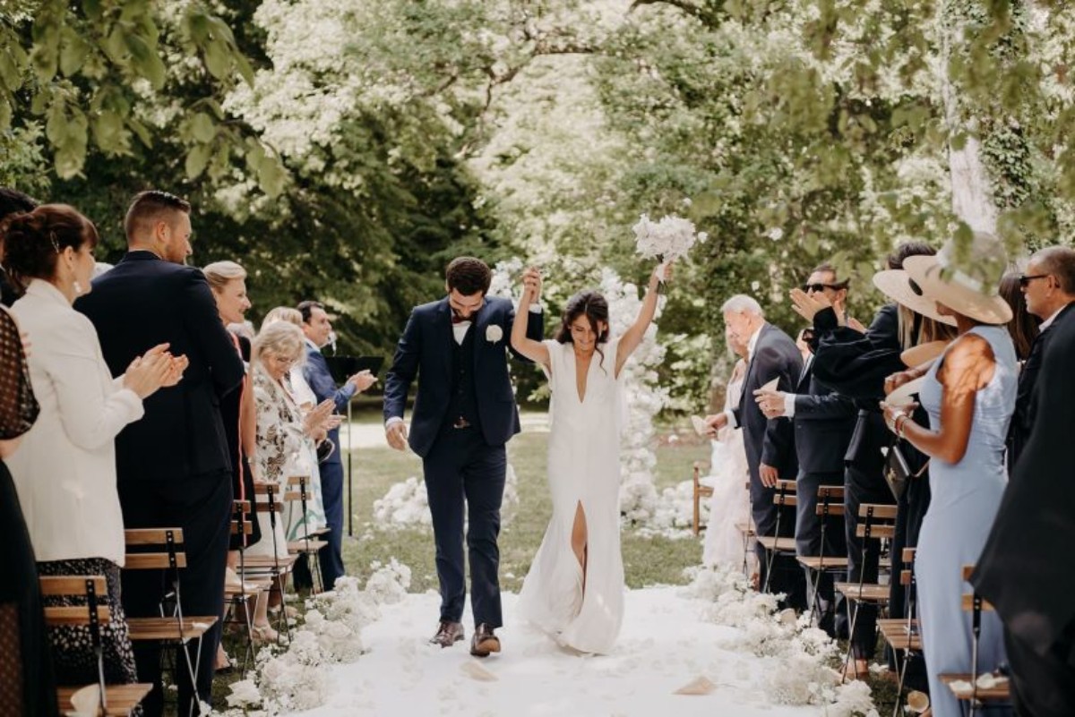 Un couple de jeunes mariés marche main dans la main après leur cérémonie de mariage en extérieur, entouré de leurs proches qui les félicitent. Une image pleine d’émotion et de joie, parfaite pour illustrer des vœux de mariage.