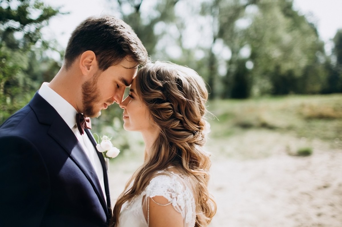 Jeune couple marié, front contre front, en extérieur, partageant un moment complice pour célébrer 1 an de mariage ; la douceur et la légèreté symboliques des noces de coton transparaissent dans cette scène romantique.