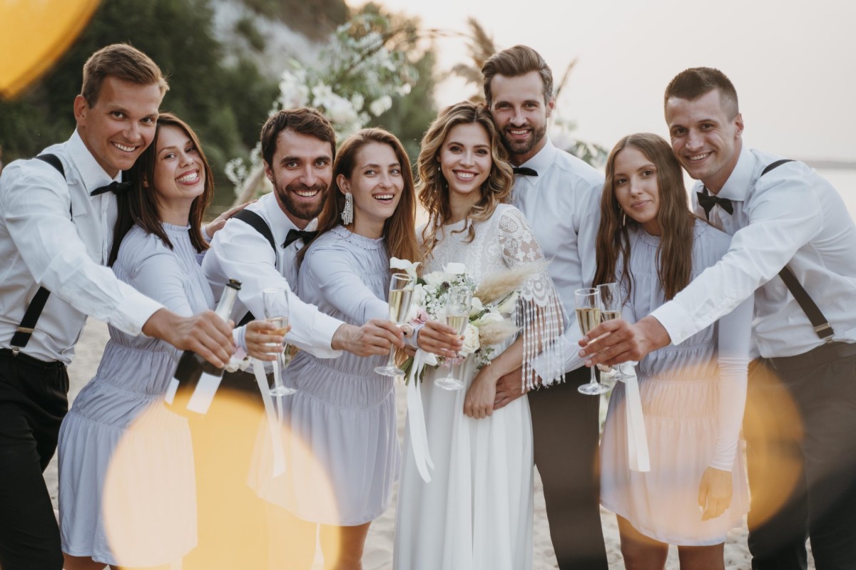 Photo de mariés entourés de leurs témoins levant des coupes de champagne lors d’une célébration de mariage sur la plage, illustrant la complicité entre les témoins et le couple.