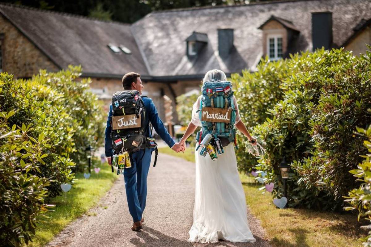 Getrouwd koppel vertrekt op huwelijksreis met rugzakken "Just Married", een origineel en avontuurlijk idee voor de huwelijksreis.